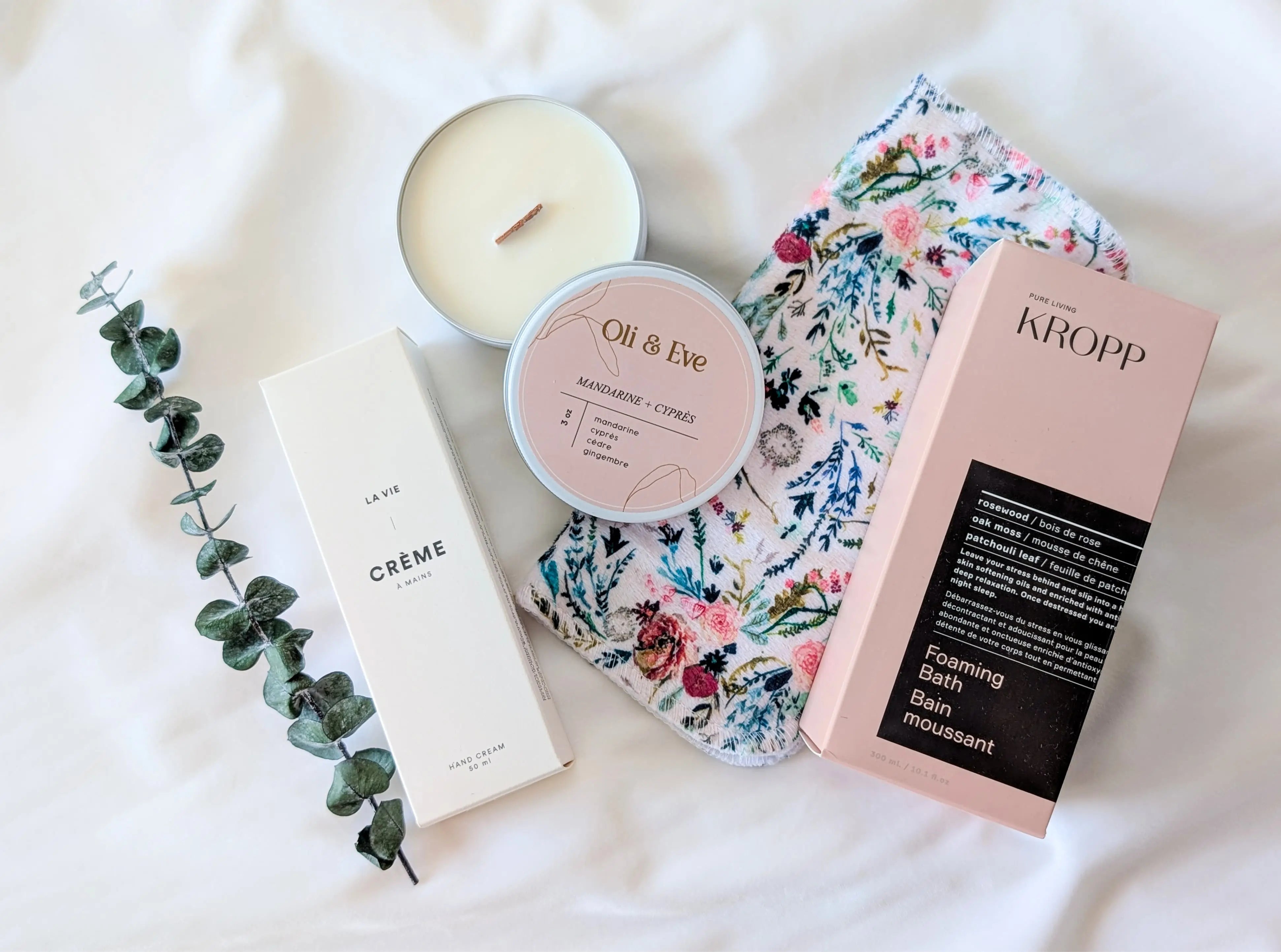 Produits de la boîte cadeau « Sérénité » disposés à plat : bain moussant Kropp aux huiles essentielles, bougie crépitante Oli & Eve, crème à mains La Vie Apothicaire, débarbouillette florale douce et branche d’eucalyptus, pour un coffret détente et bien-être québécois.